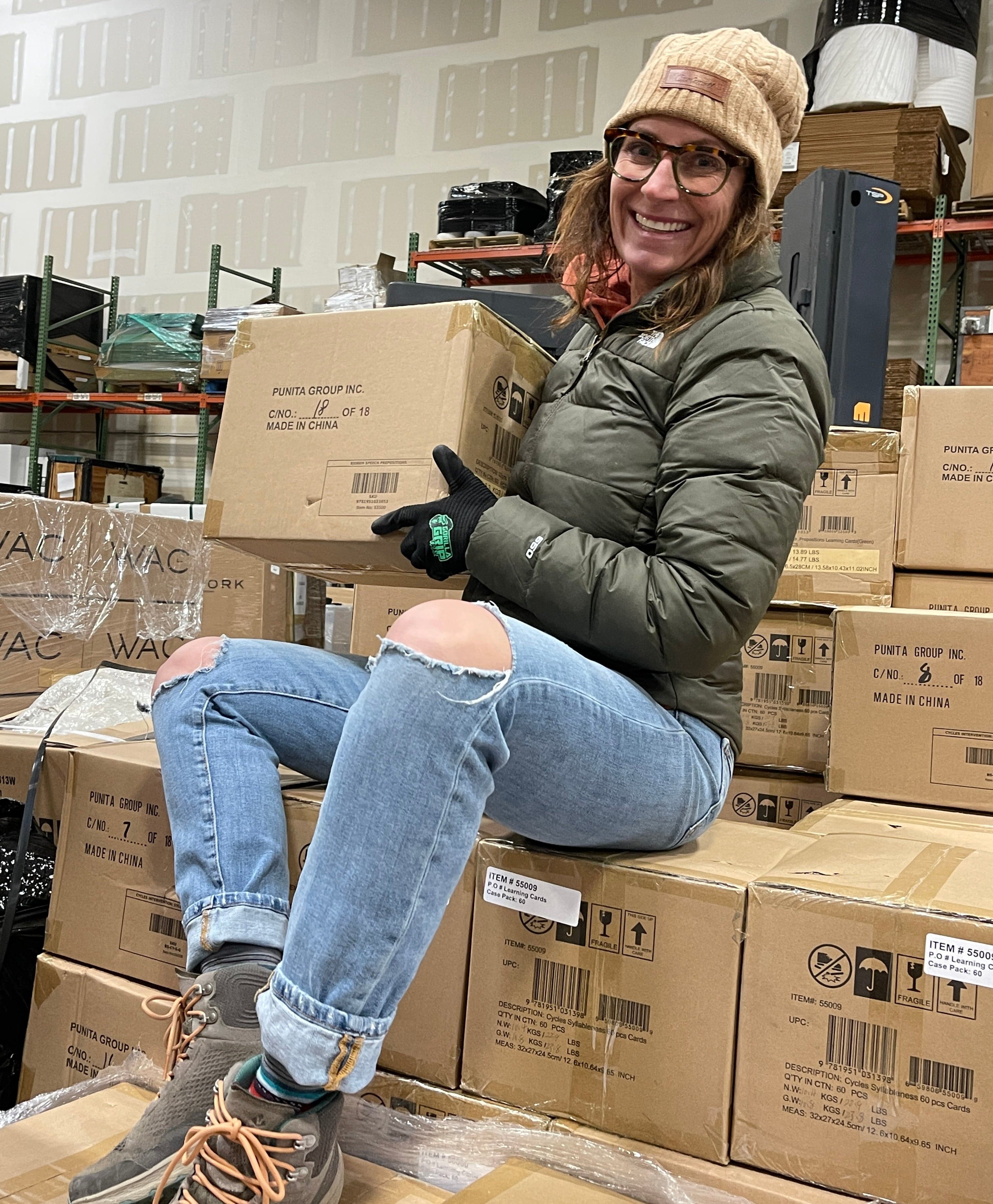 Woman in beanie and jacket sitting on boxes, holding a box.