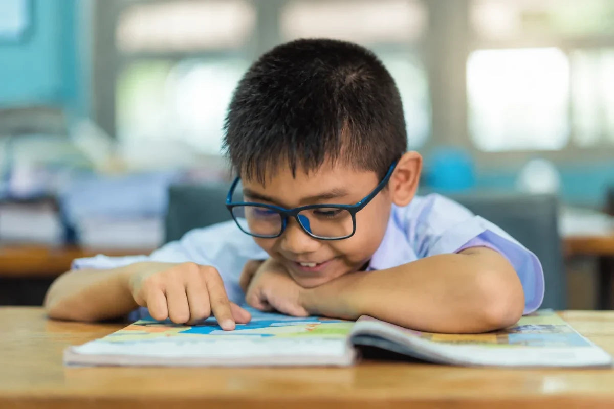 Boy in glasses reading a book at a classroom desk